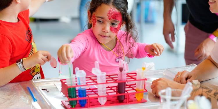 A child with facepaint on using a pipette to move different coloured liquids between test tubes.