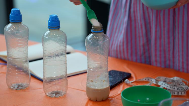A teaspoon of sugar is being poured into a clear plastic bottle containing 5 centimetres of a brown liquid