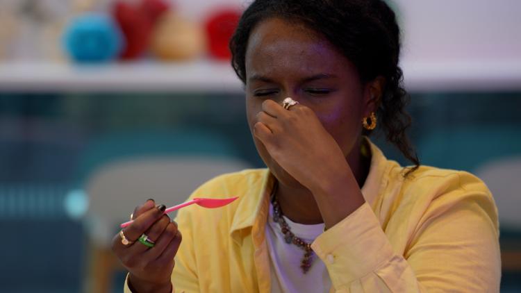 A woman who has black hair and is wearing a yellow shirt has her eyes closed, is pinching her nose and is holding a spoon. She is frowning.