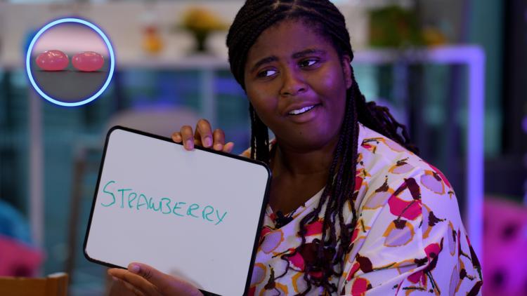 A smiling woman wearing a floral top holds up a small whiteboard with 'strawberry' written on it. An image of 2 pink jellybeans is overlayed in the top left corner.