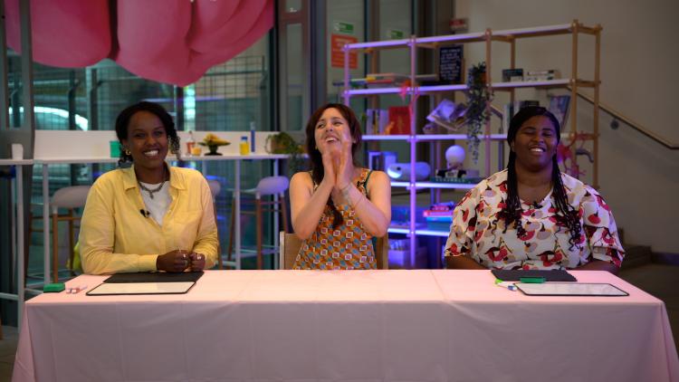 Three women sit behind a table with whiteboards in front of them. They are all laughing and the woman in the middle is clapping.