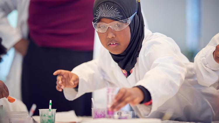 A child wearing safey glasses and a lab coat with different coloured pots of glitter.