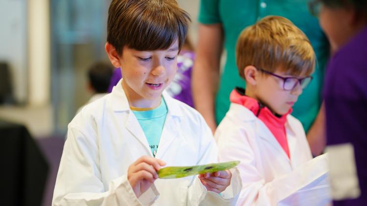 Two kids in lab coats and safety glasses holding pieces of paper.