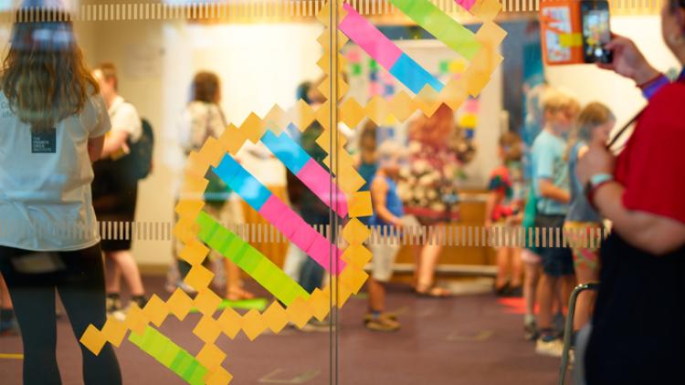 A DNA double helix on a window made out of post it notes, with people either side of the glass.