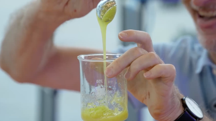 Someone holding a clear glass beaker filled with green liquid, stirring it with a spoon.