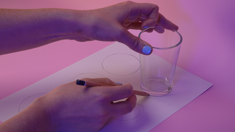 Someone holds a drinking glass in one hand and a pencil in the other hand. They are drawing around the glass to draw a perfect circle on a piece of paper