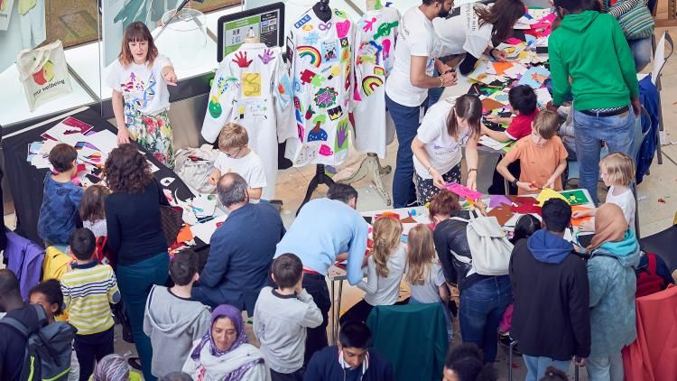 A crowd of people decorating two hung-up labcoats which are covered in colourful patches.