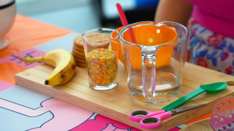A banana, biscuits, a cup of sprinkles, and plastic bowl and spoon, a measuring jug containing water, scissors, a spoon and a masher are laid out on a wooden chopping board.