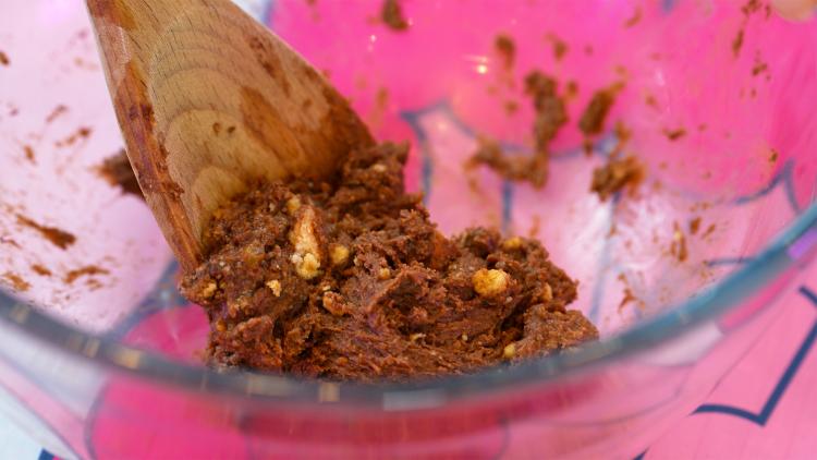 A brown mixture with chunks of biscuit is being mixed with a wooden spoon