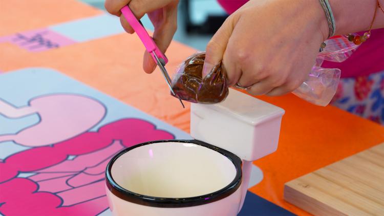 Above a toilet-shaped mug, someone uses scissors to snip a hole in a plastic sandwich bag which contains a brown mixture