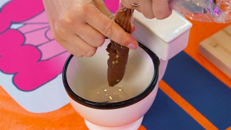 A hand squeezes a brown mixture out of a plastic sandwich bag. The mixture takes the shape of a fake poo.