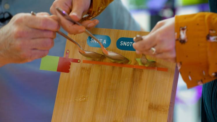 People holding spoons of green liquid against a wooden chopping board, so that the liquid does not spill down the chopping board.