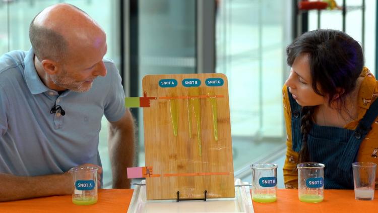Two people look at a propped up wooden chopping board with three streams of green liquid running down it.