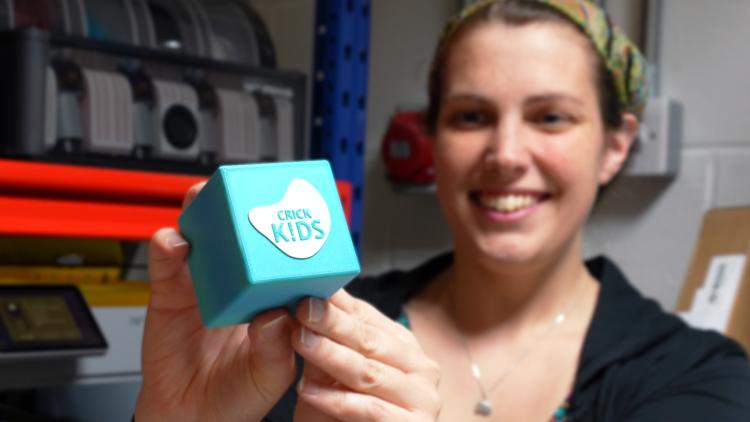 Smiling woman holding a 3D-printed, turquoise-coloured cube with the words 'Crick Kids' printed onto it